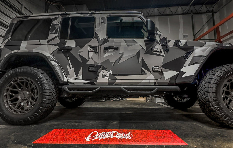 Camouflage-patterned SUV in a garage with a red 'Paint Dead' mat on the floor.
